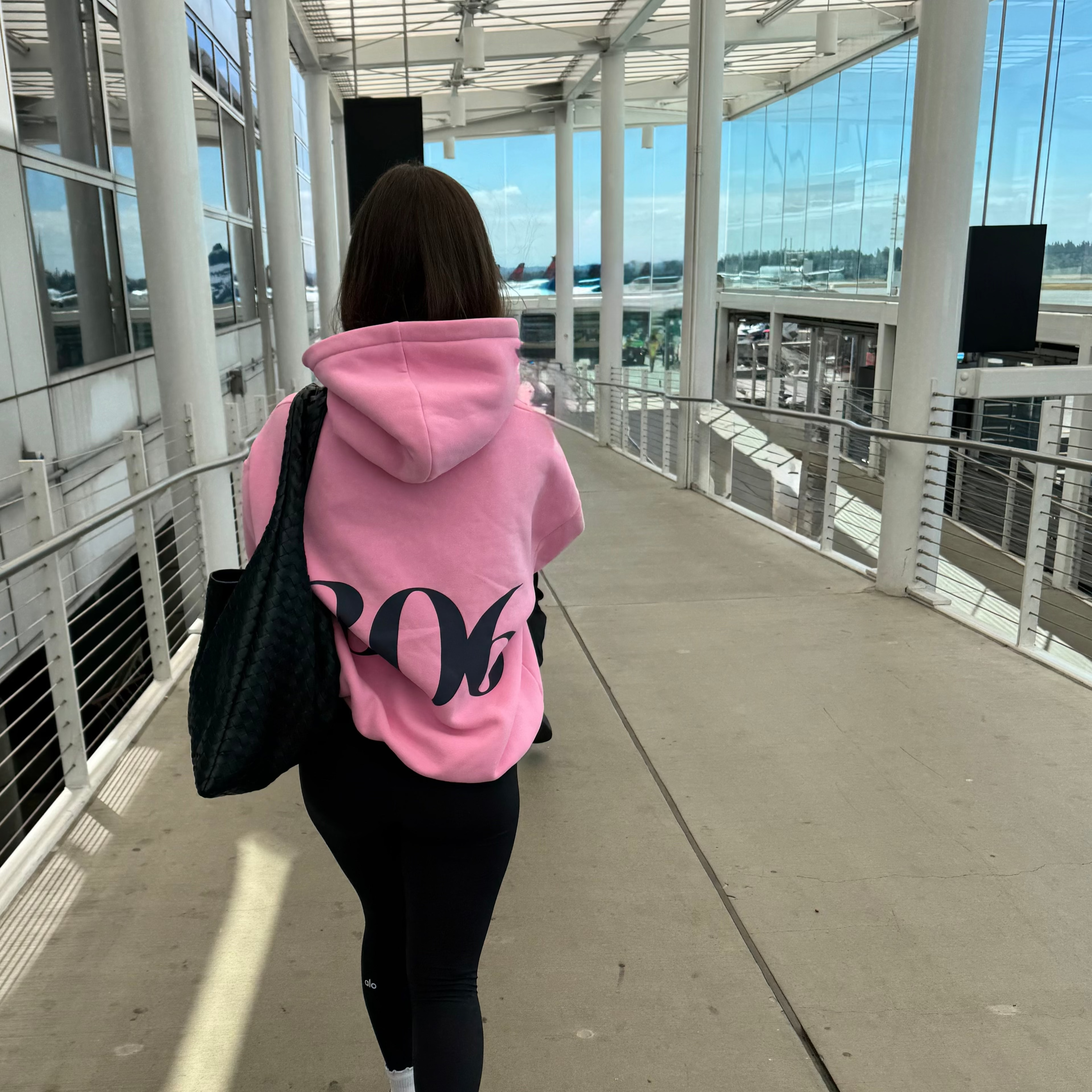 Person wearing a pink hoodie with '306' printed on it, walking in an airport terminal.